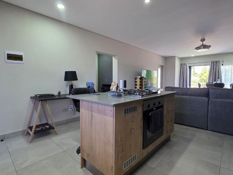 Modern open-plan living area featuring a central kitchen island with built-in oven and gas hob, complemented by neutral tiled flooring and recessed lighting.