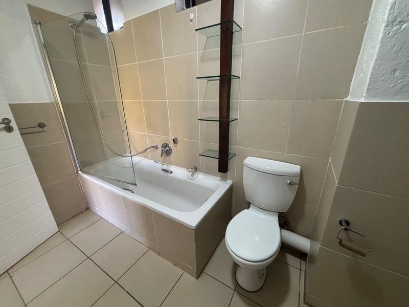A functional bathroom featuring a bathtub with a glass shower screen, a standard toilet, and neutral wall and floor tiling.