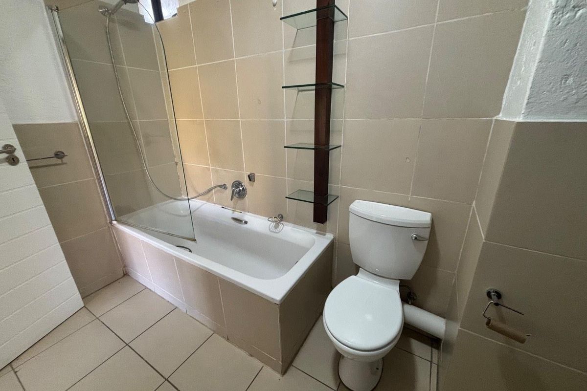 A functional bathroom featuring a bathtub with a glass shower screen, a standard toilet, and neutral wall and floor tiling.