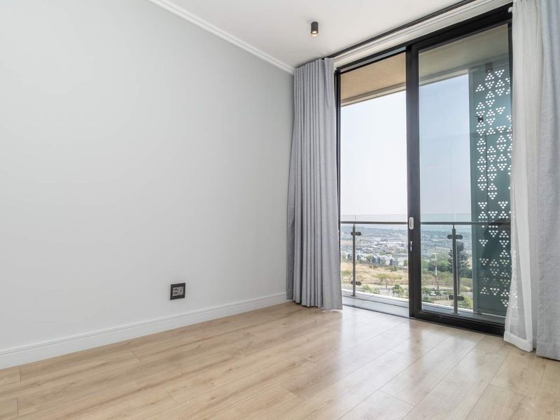 A bright, empty bedroom featuring light wood-look laminate flooring, modern recessed lighting, and a large sliding glass door opening to a balcony with scenic views.