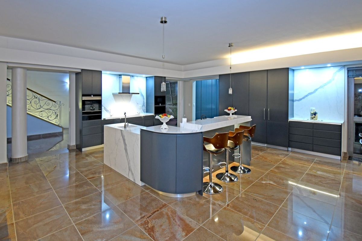 Modern kitchen featuring dark cabinetry, stone countertops, a central island with breakfast bar, built-in appliances, and polished tiled flooring.