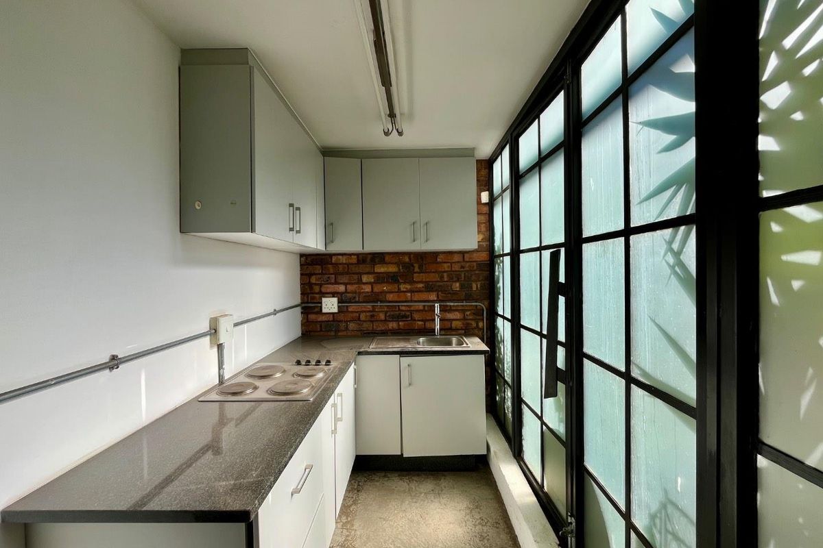 Compact kitchenette featuring built-in cabinetry, stone-look countertops, integrated electric hob, stainless steel sink, and a distinctive exposed brick feature wall.