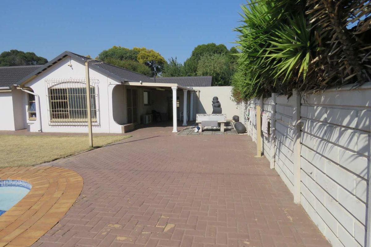 Rear exterior view featuring a paved patio area, covered veranda with columns, swimming pool edge, and perimeter boundary wall.