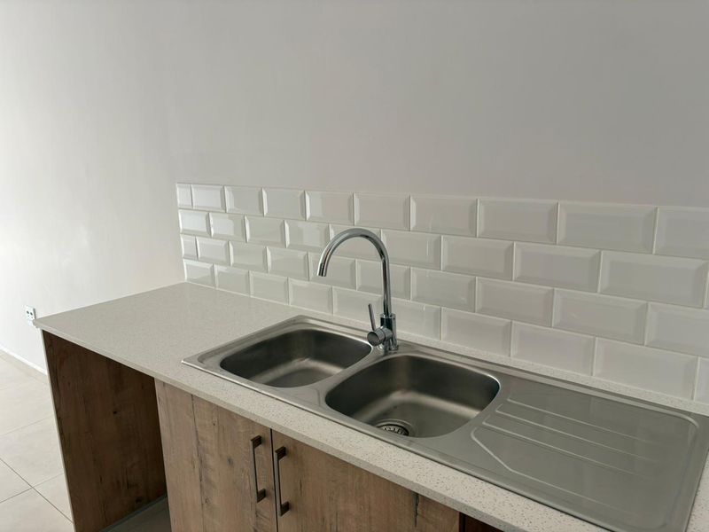 A clean, modern kitchen area featuring a stainless steel double sink, chrome mixer tap, subway tile backsplash, and wood-grain cabinetry with stone-look countertops.