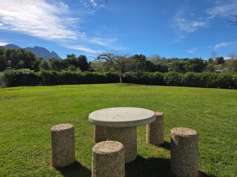 A well-maintained private garden featuring a lush green lawn, mature hedging, and a permanent concrete outdoor seating area with mountain views.