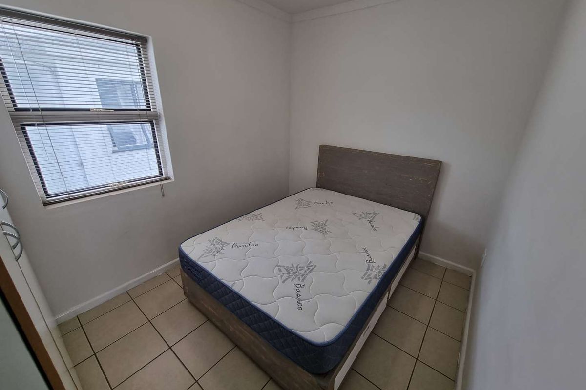 A compact bedroom featuring tiled flooring, a window with fitted blinds, and a built-in cupboard, providing a functional and neutral living space.