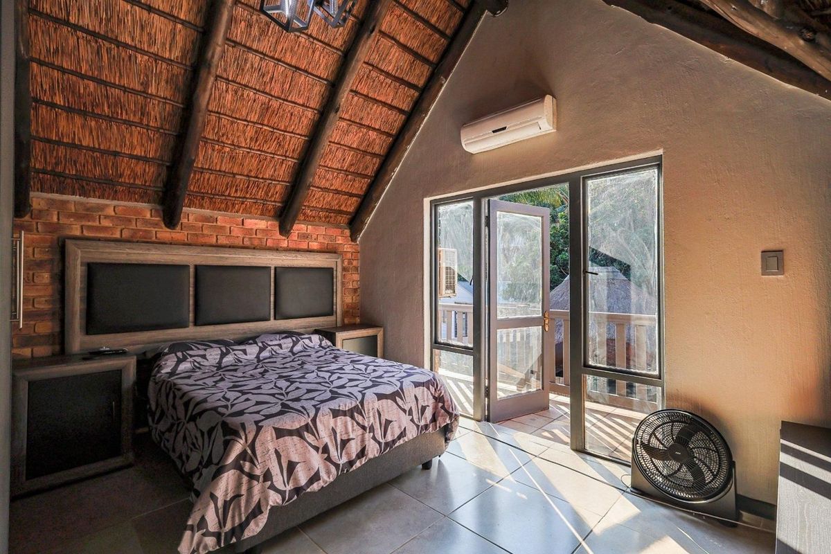 A rustic bedroom featuring a high thatched ceiling with exposed timber rafters, brick feature wall, tiled floors, and direct access to a private balcony.