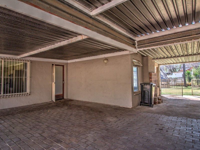 A covered outdoor parking area or garage featuring paved flooring, corrugated metal roofing, and security bars on the windows.