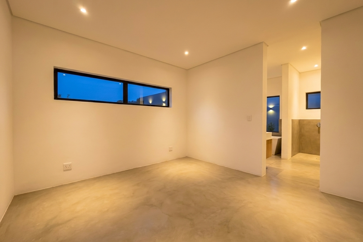 A modern, minimalist bedroom featuring polished concrete flooring, recessed ceiling lighting, and an open-plan layout leading to an en-suite bathroom area.