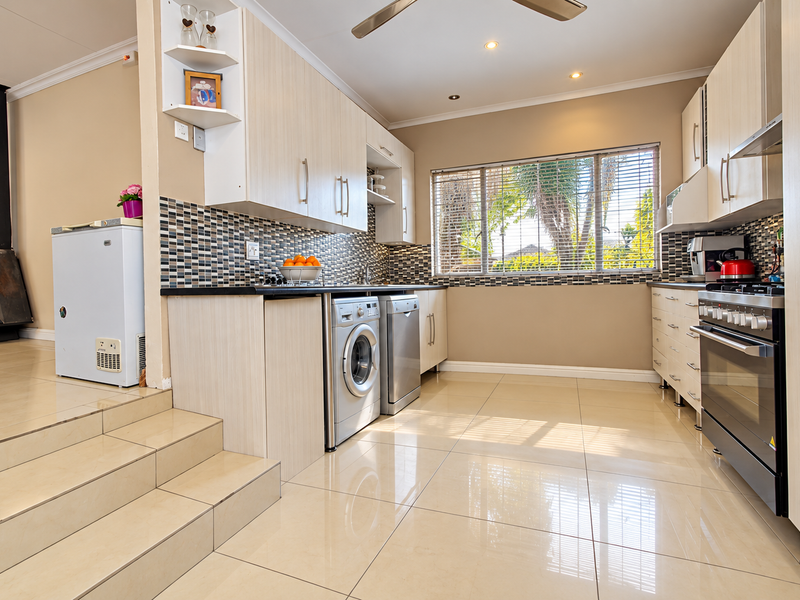 Modern kitchen featuring light wood cabinetry, mosaic tile backsplash, gas stove, and integrated appliances, with an adjacent raised living area and built-in fireplace.