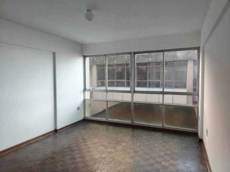 An empty living room featuring original parquet flooring and large windows providing natural light and an urban outlook.