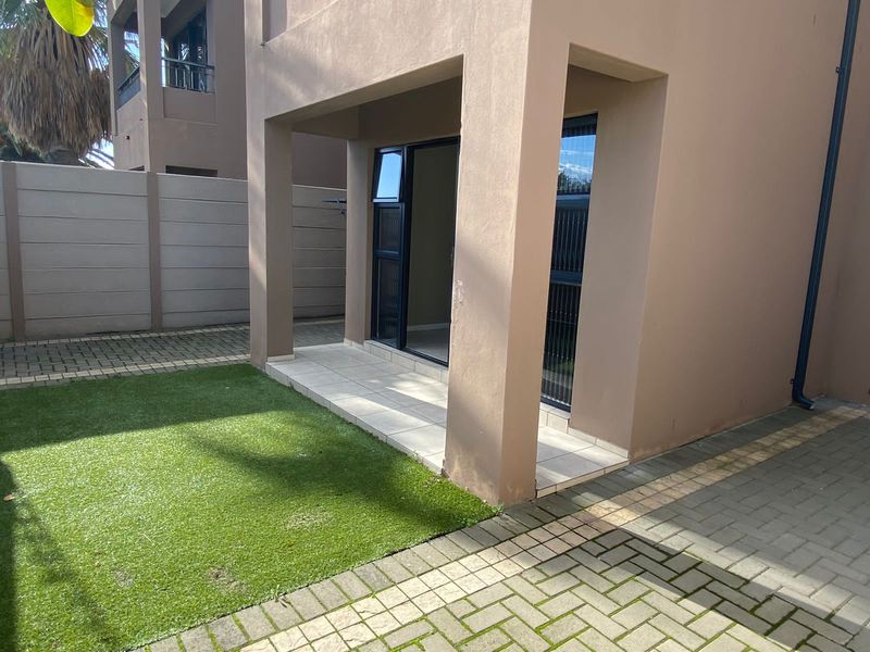 Rear exterior view featuring a covered patio with tiled flooring, artificial grass lawn, and paved area, enclosed by a precast concrete boundary wall.