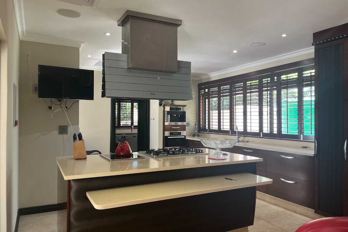Modern kitchen featuring a central island with a built-in gas hob, overhead extractor fan, dark cabinetry, and plantation shutters on the windows.