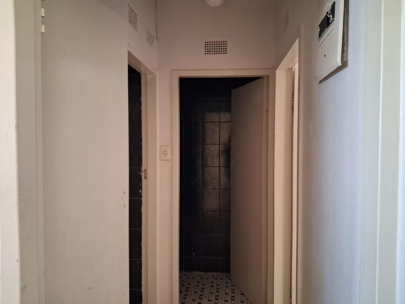 A narrow apartment hallway featuring plain walls, ventilation grilles, and access to adjacent rooms with dark tiled flooring visible.