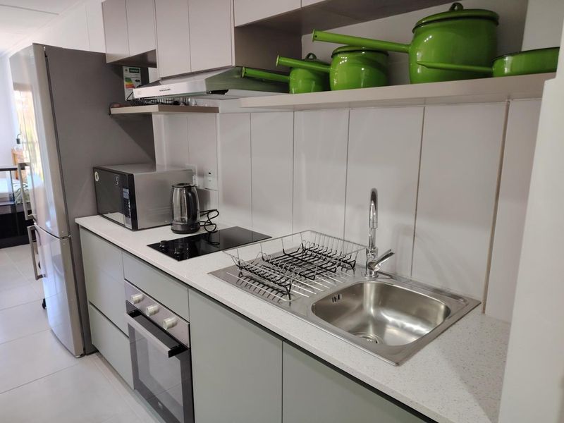 A modern, compact kitchen featuring light grey cabinetry, stone countertops, a stainless steel sink, and integrated appliances including an oven and electric hob.
