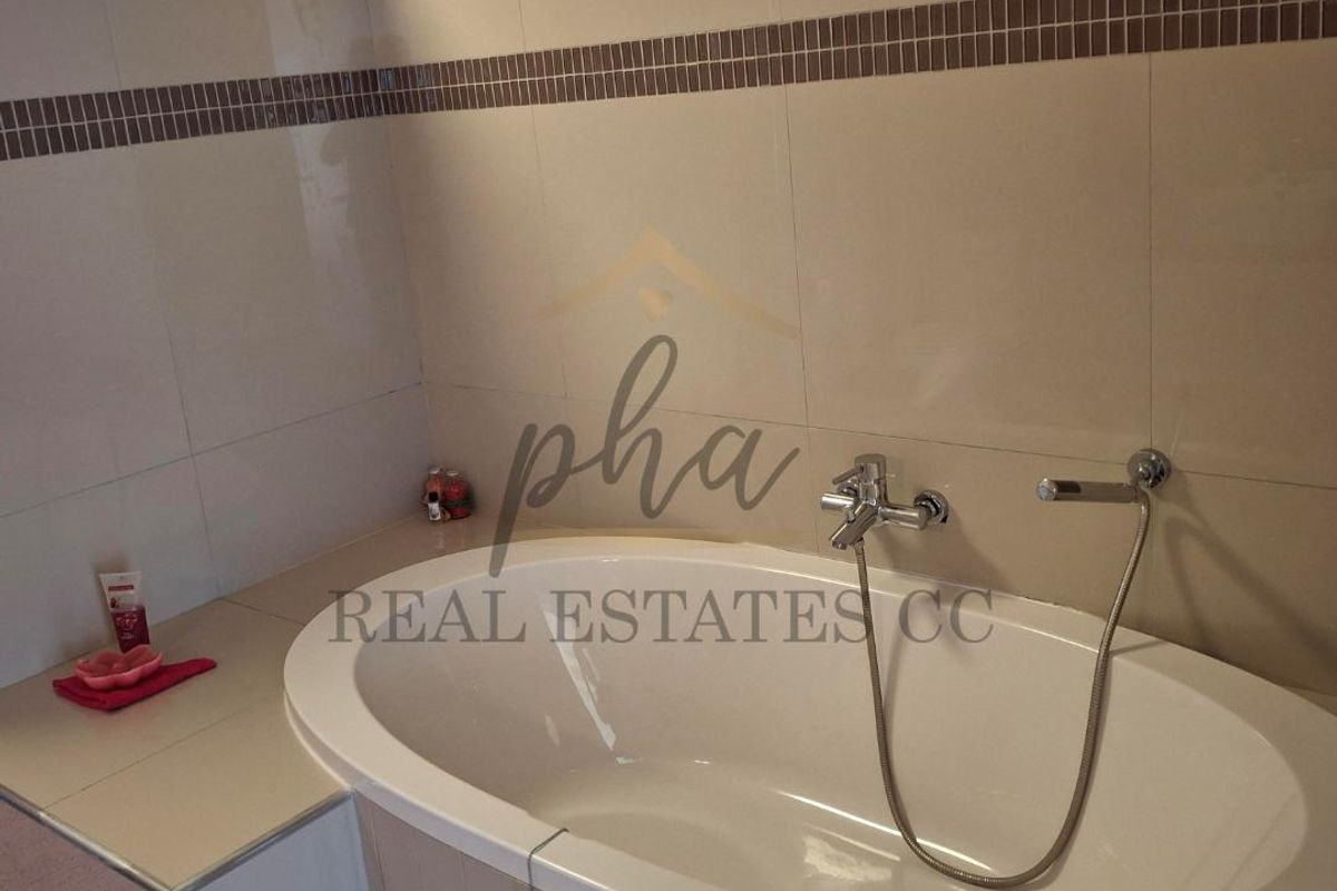 A modern bathroom featuring a built-in bathtub with chrome wall-mounted faucet and shower head, complemented by neutral wall tiling and a decorative mosaic border.