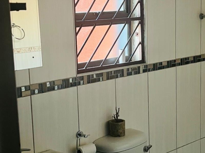 A functional bathroom featuring neutral ceramic wall tiles, a pedestal basin, and a toilet with security bars on the window.