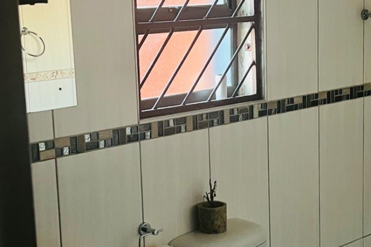 A functional bathroom featuring neutral ceramic wall tiles, a pedestal basin, and a toilet with security bars on the window.