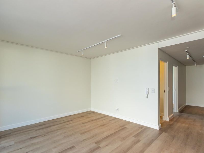 A bright, empty living area featuring modern laminate flooring, clean white walls, and contemporary track lighting, leading into a hallway.