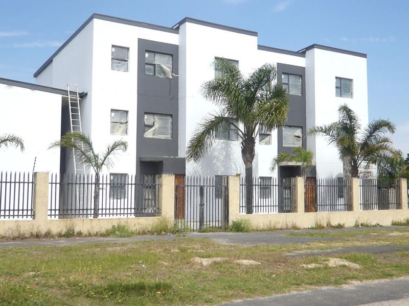 Front elevation of a multi-story apartment complex featuring modern white and grey facade finishes, secured by perimeter fencing and a gated entrance.