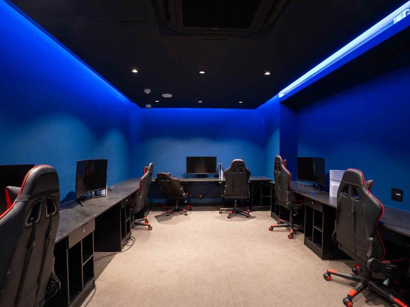 A modern, dedicated gaming or media room featuring built-in desk workstations, ambient cove lighting, and recessed ceiling fixtures.