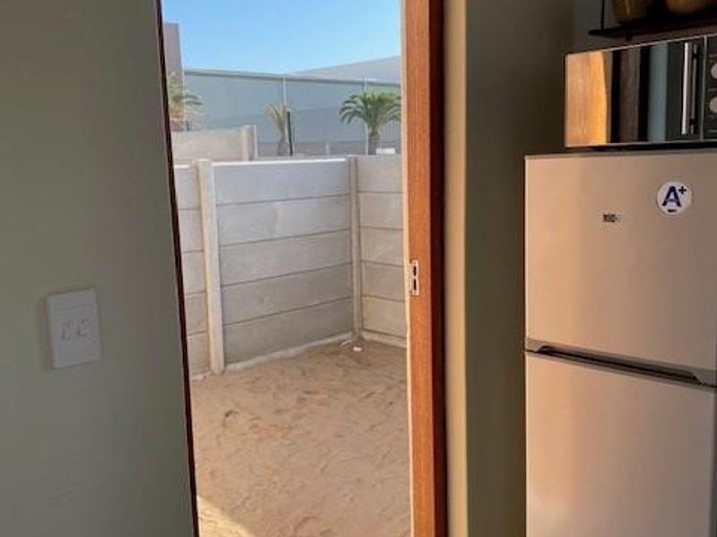 A kitchen area featuring tiled flooring and a timber door leading out to an unlandscaped sandy rear yard with a precast concrete boundary wall.