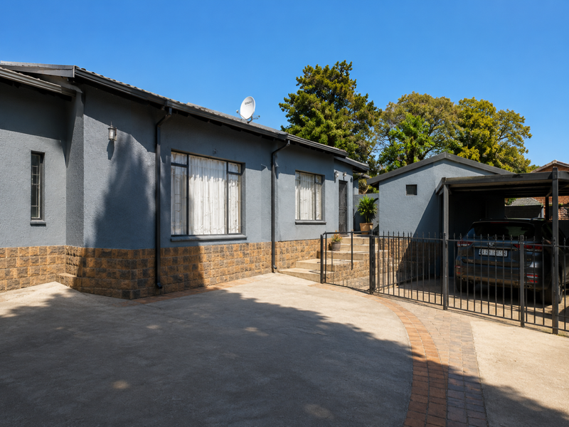 Front exterior view of the house featuring a concrete driveway, stone-clad base, and a covered carport with metal security fencing.