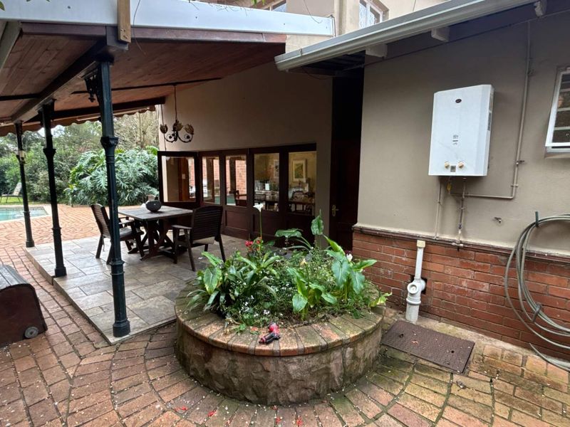A covered outdoor patio area featuring brick paving, a central brick planter, and a wall-mounted gas geyser, overlooking the garden and pool area.
