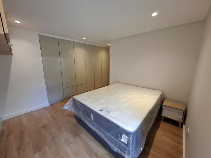 A modern bedroom featuring light wood-look laminate flooring, recessed ceiling lighting, and extensive floor-to-ceiling built-in cupboards with a neutral finish.