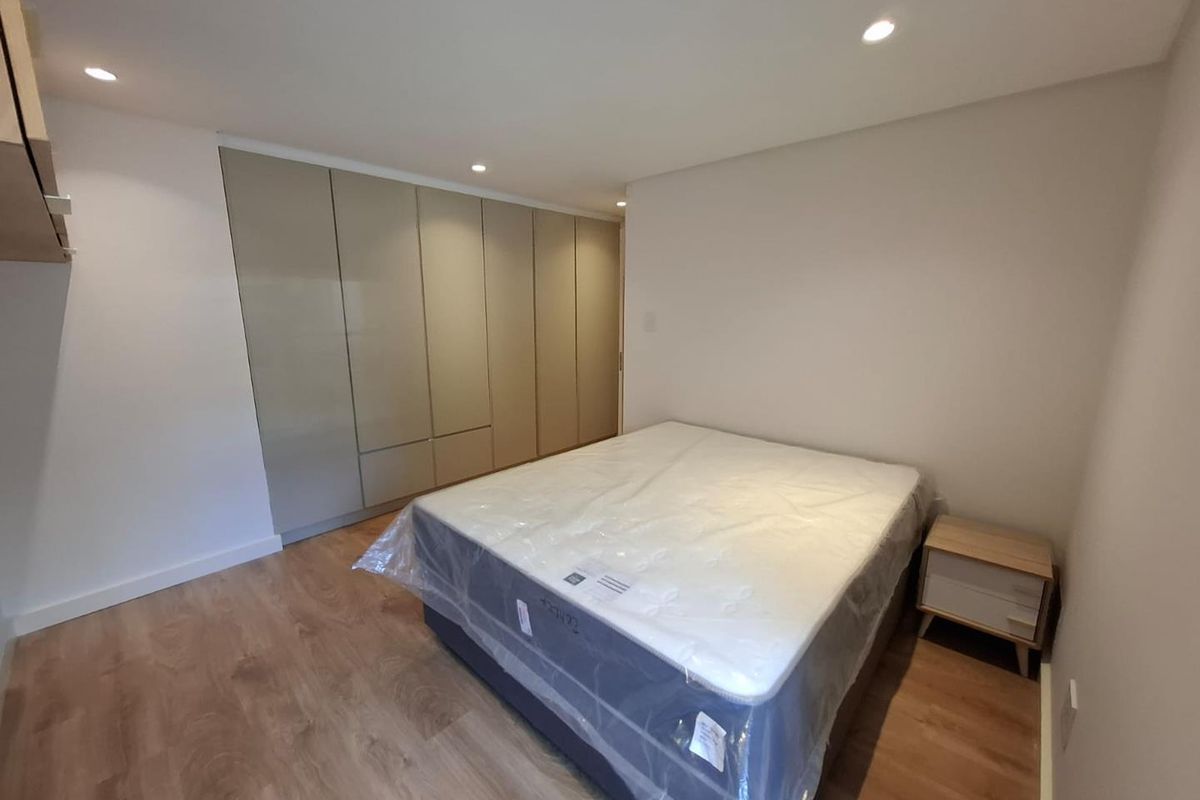 A modern bedroom featuring light wood-look laminate flooring, recessed ceiling lighting, and extensive floor-to-ceiling built-in cupboards with a neutral finish.