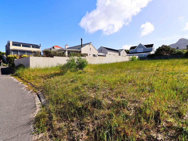 A vacant residential plot of land situated on a paved street, featuring natural grass coverage and an existing boundary wall.