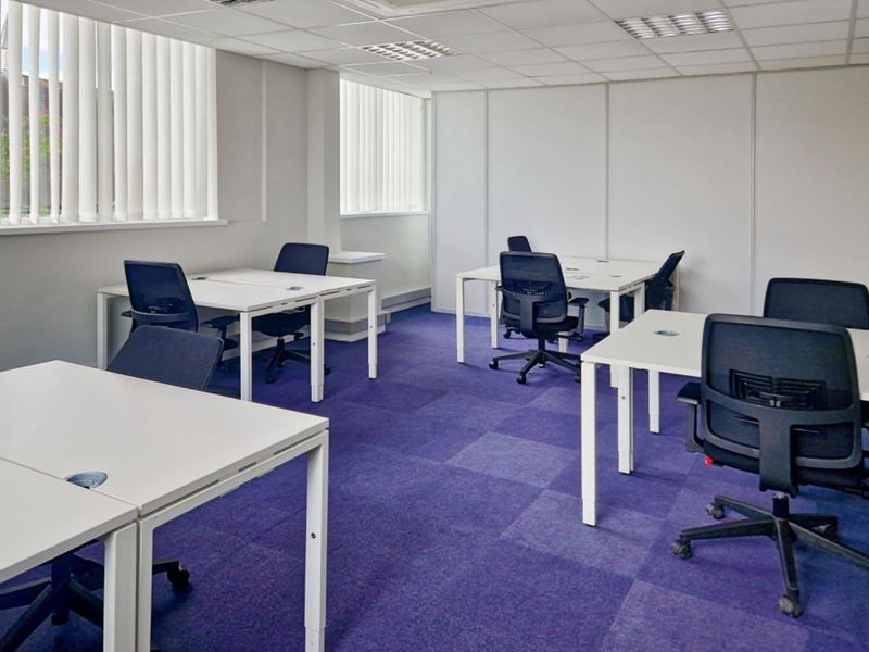 A functional office space featuring a suspended ceiling, recessed lighting, vertical window blinds, and durable carpet tiles.