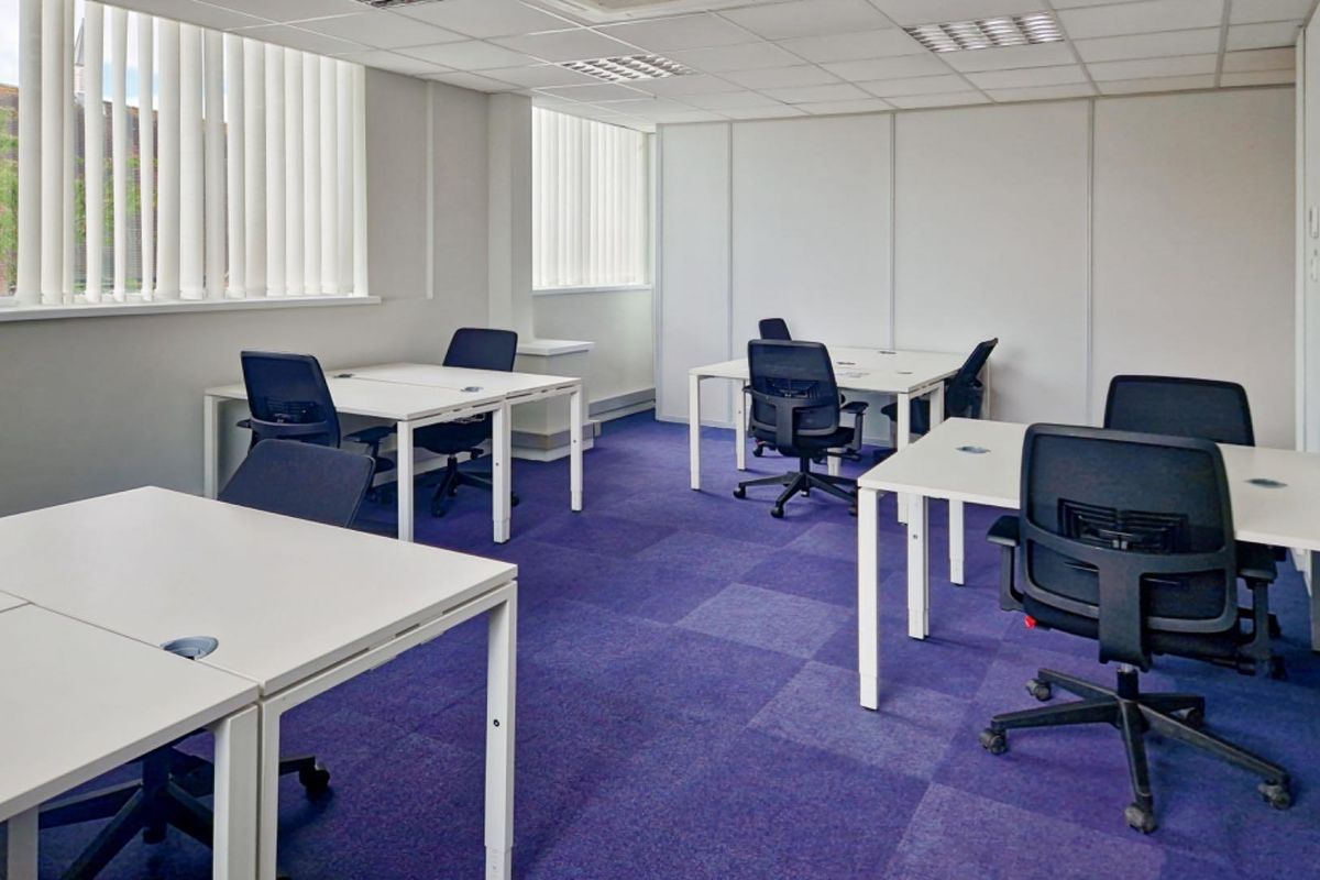 A functional office space featuring a suspended ceiling, recessed lighting, vertical window blinds, and durable carpet tiles.