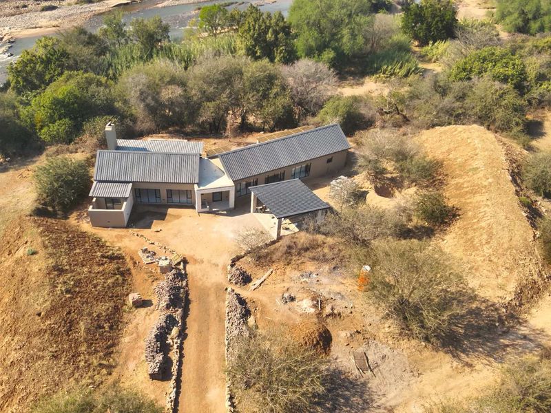 Aerial view of a modern house with a metal roof, covered patio, and driveway, situated in a natural landscape near a river.