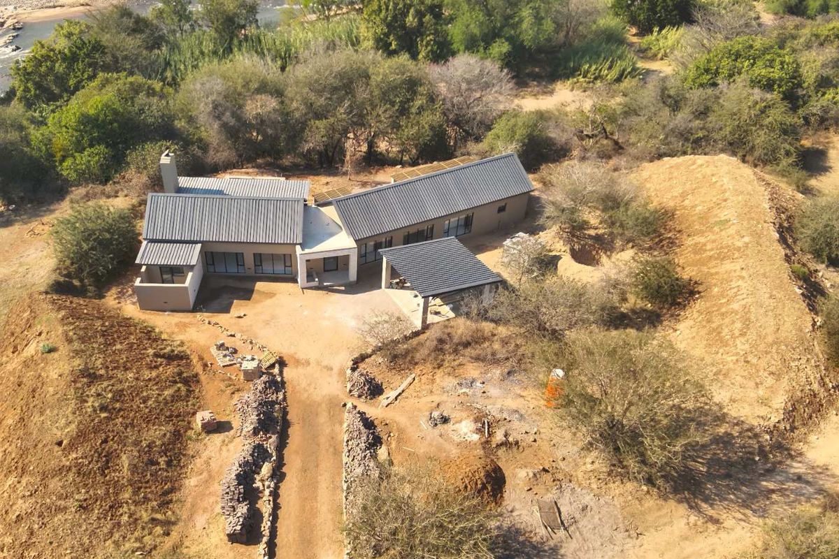 Aerial view of a modern house with a metal roof, covered patio, and driveway, situated in a natural landscape near a river.