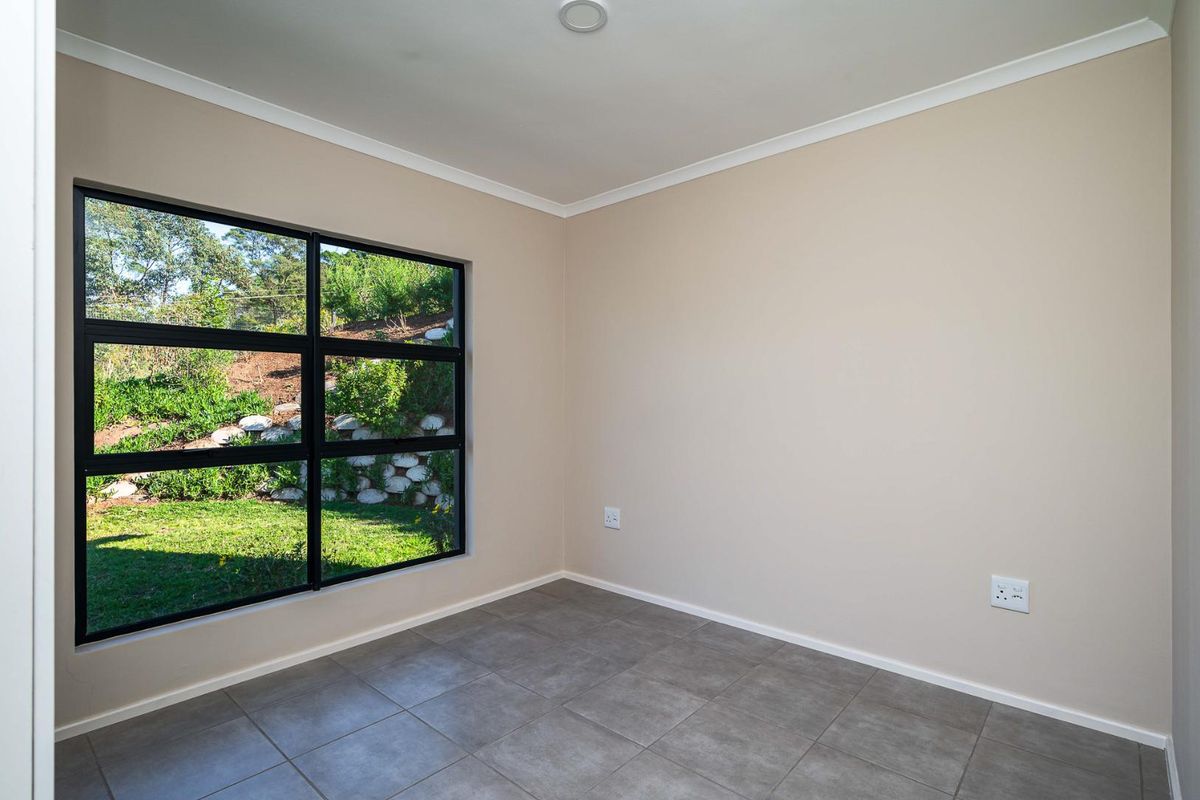 A bright, empty bedroom featuring neutral wall tones, large black-framed windows overlooking the garden, and modern grey floor tiles.