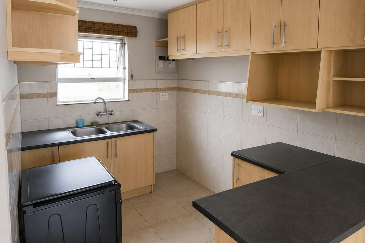 A functional kitchen featuring light wood cabinetry, dark countertops, tiled splashbacks, and a double stainless steel sink with a window view.