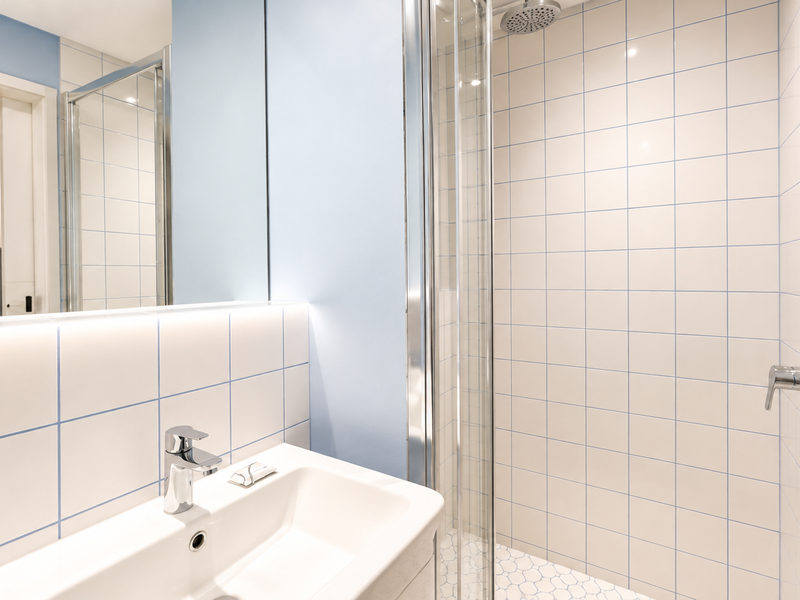 A clean, modern bathroom featuring a glass-enclosed shower, white tiled walls with blue grout, a vanity basin, and built-in storage shelving.