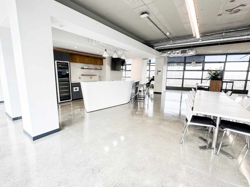 Modern open-plan office or commercial space featuring polished concrete floors, an industrial exposed ceiling, a built-in bar area, and expansive windows with natural light.