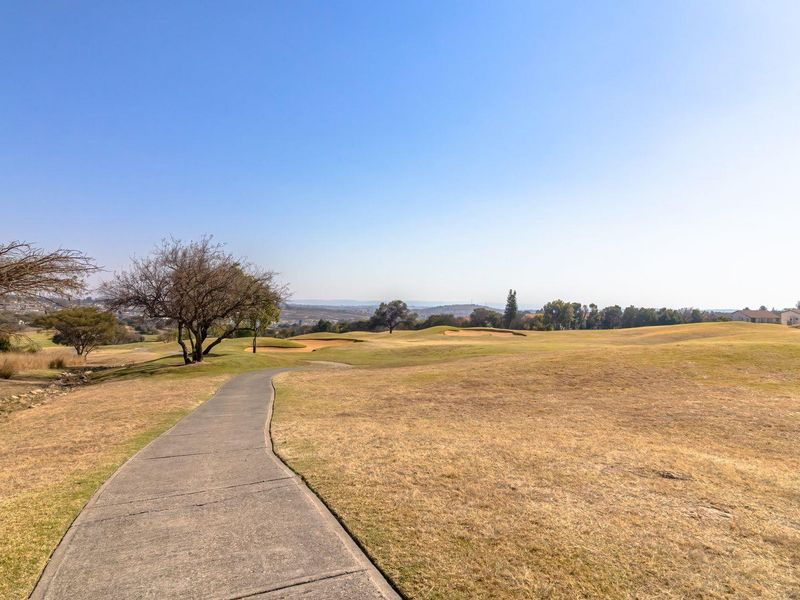 A scenic view of a golf course landscape featuring a paved pathway winding through the open, grassy terrain under a clear blue sky.