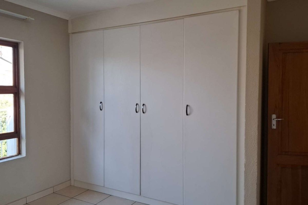 A clean, empty bedroom featuring neutral walls, tiled flooring, and ample built-in cupboard storage with a window providing natural light.