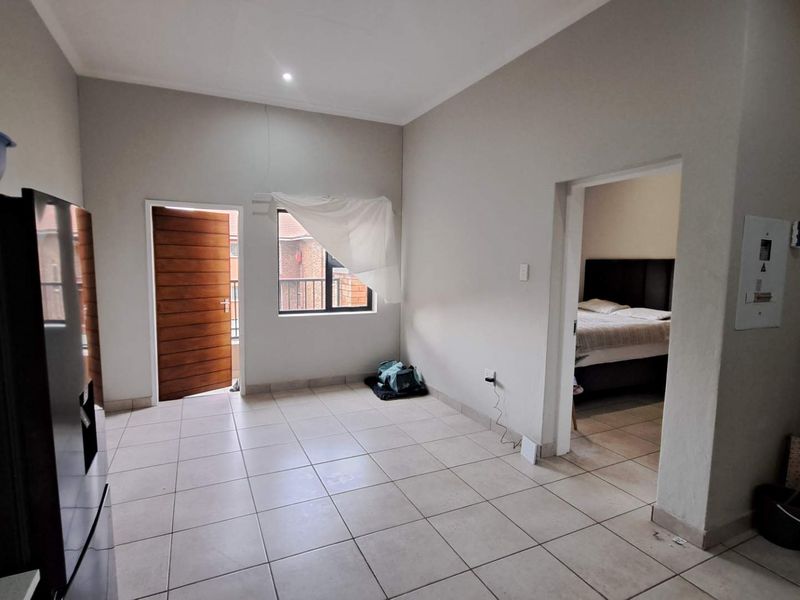 An open-plan living area featuring neutral tiled flooring, a wooden exterior door, and direct access to an adjacent bedroom.