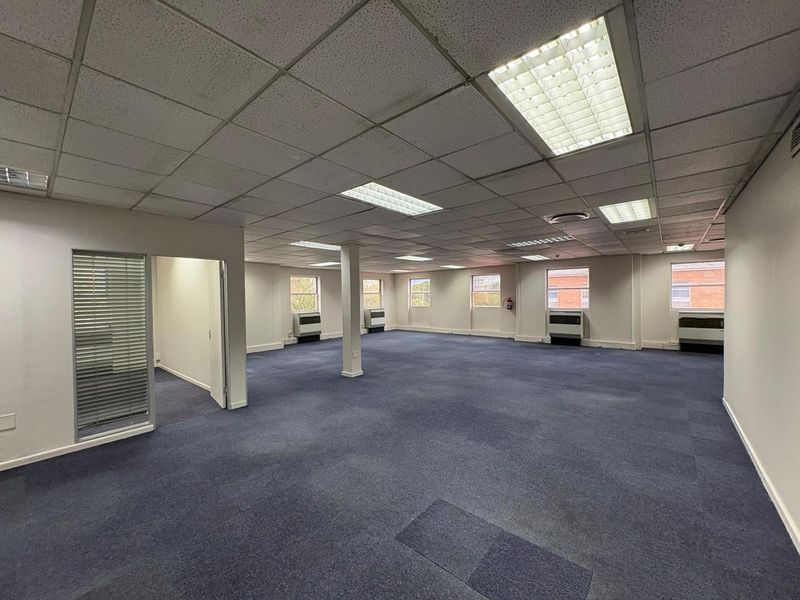 Spacious open-plan office area featuring suspended ceilings, recessed lighting, carpeted flooring, and multiple window-mounted air conditioning units.