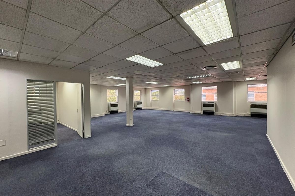 Spacious open-plan office area featuring suspended ceilings, recessed lighting, carpeted flooring, and multiple window-mounted air conditioning units.