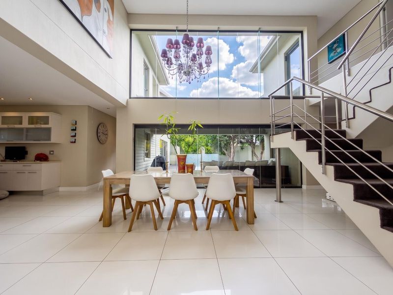 Spacious open-plan dining area featuring high ceilings, large glass windows, modern staircase with stainless steel railing, and sleek tiled flooring.