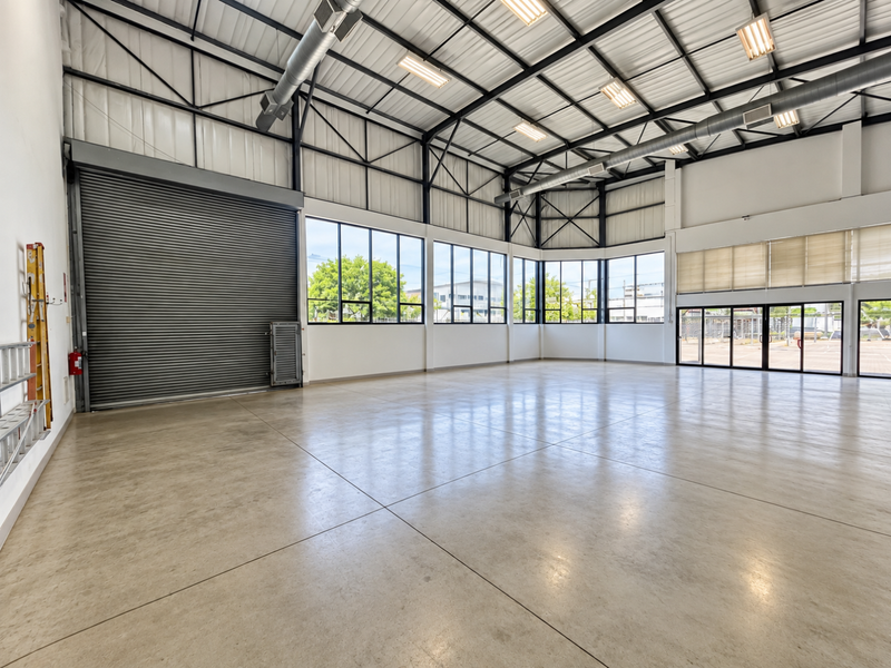 Spacious industrial utility space featuring polished concrete floors, high exposed steel truss ceilings, large windows, and a secure roller shutter door.