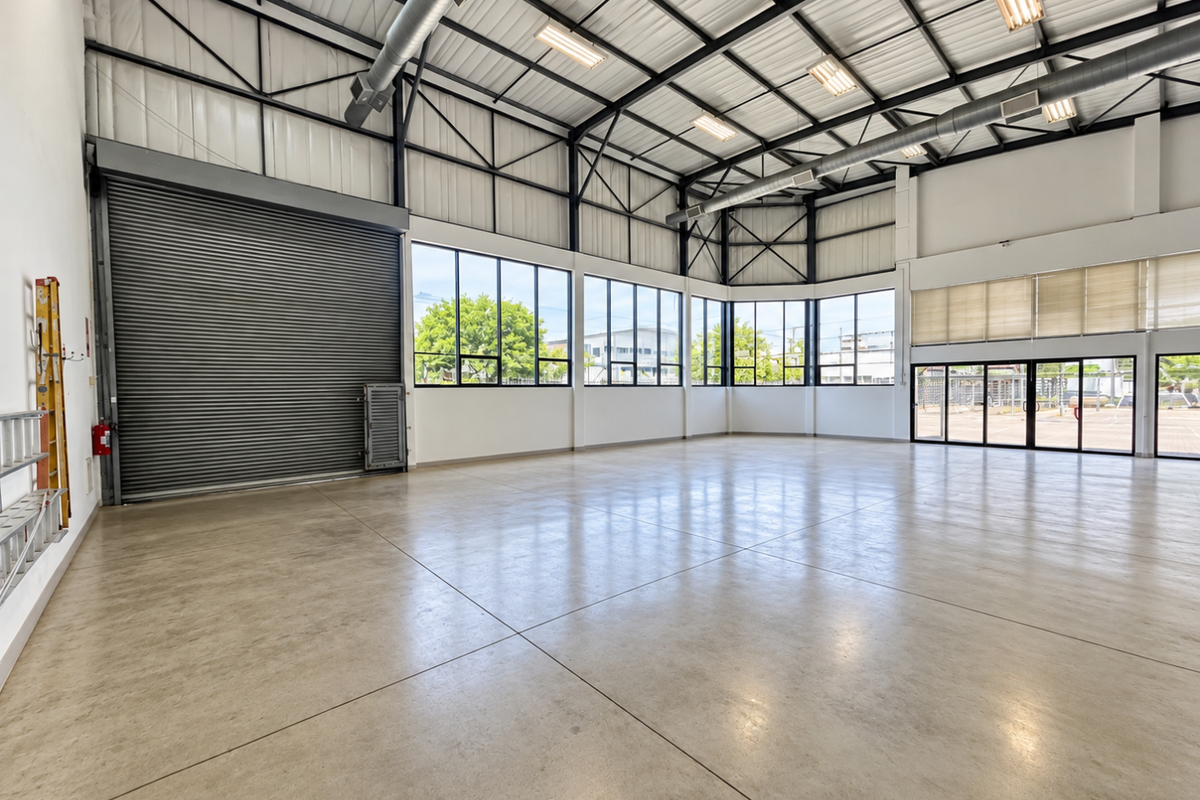 Spacious industrial utility space featuring polished concrete floors, high exposed steel truss ceilings, large windows, and a secure roller shutter door.