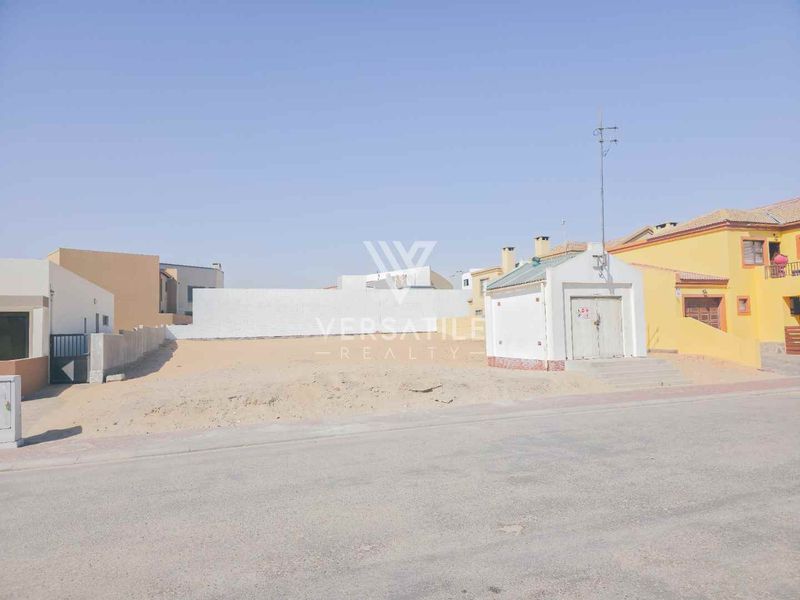 A vacant residential plot featuring a boundary wall and a small utility structure, situated along a paved street in a residential neighborhood.