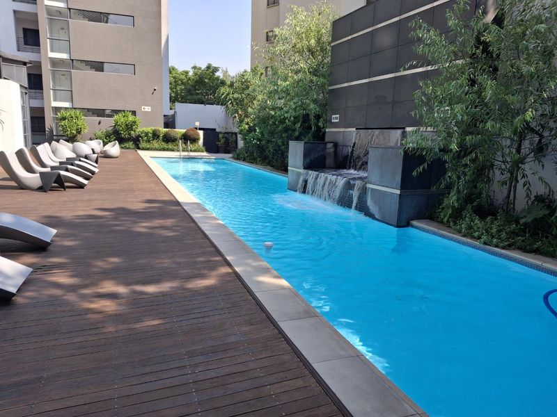 A well-maintained communal swimming pool area featuring a modern water feature, stone cladding, and timber decking in a contemporary apartment complex setting.