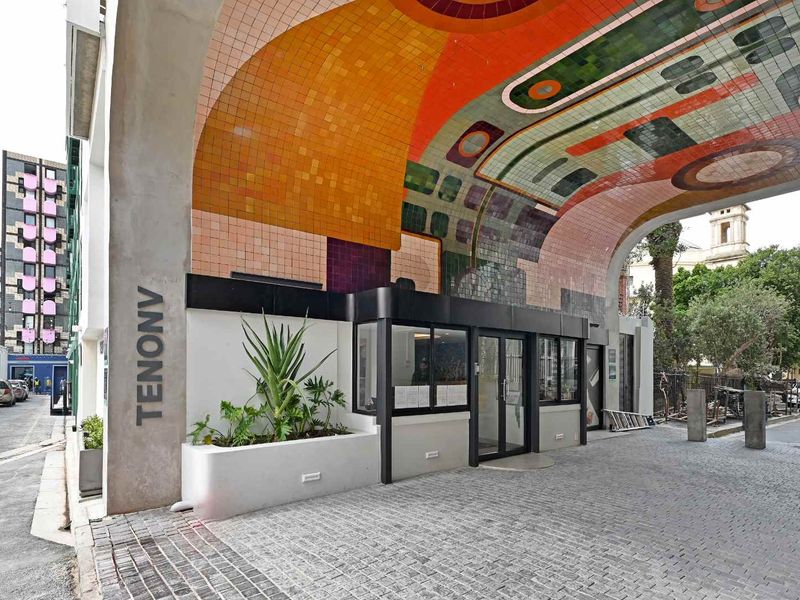The front entrance of the apartment building features a striking, colorful tiled archway, a secure glass-fronted lobby entrance, and a paved cobblestone driveway.